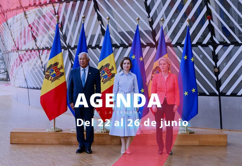 Tres personas de pie frente a banderas en un evento oficial.